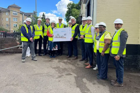 Jaimielee Rendall/BBC A group of men and women all wearing hard hats and yellow high-vis jackets. They are standing in front of a former pub building. They are smiling. Two of them are holding up an artist's impression image of what the pub will look like after its conversion.