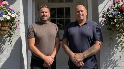 Martyn Pope Two men smiling at the camera outside a white building. The one on the left has a brown t-shirt on and short brown hair and has his arms in front of him with his hands clasped together. The one on the right is bald with a blue t-shirt on, with his arms in front of him and his hands clasped together.