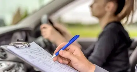 Getty Images A driving tester is in the passenger seat of a car, assessing a new driver as they take their test. In the foreground the tester is marking a document on a clipboard and blurred in the background the driver can be seen at the wheel.