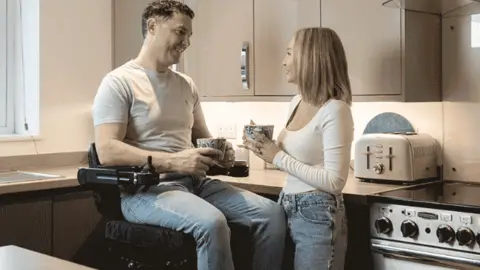 Conquering Horizons Man in a powered wheelchair. It lifts off the ground and is adjustable in height. There's a chap in a grey T-shirt and jeans. A women is stood next to him at the same height level in the kitchen holding a mug in her hand.