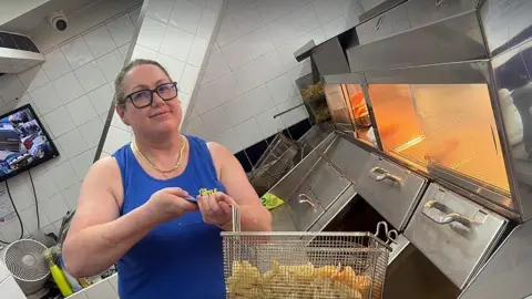The Little Chippy Anna Leigh with brown hair tied back in a pony tail wearing black glasses and a blue sleeveless t-shirt stands behind the frying range in a fish and chip shop. She holds up a frying basket of chips. She is smiling.