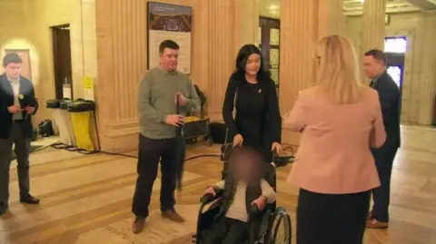 A photo from the event at Stormont showing Michael McMonagle standing to the left of the screen looking down at his phone, he is wearing trousers, shirt and a jacket. Michelle O'Neill stands to the right of the screen with her back to the camera, wearing a pink jacket. In the centre of the image is a child in a wheelchair with their face blurred and their parents standing behind him.
