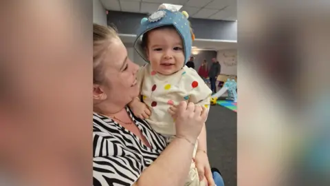 A little boy looking at the camera and smiling. He is wearing a blue birthday crown with a "1" badge pinned on it. He is sat on a woman's knee and she is looking at him and smiling. There is play equipment in the background.