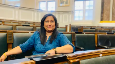 Birmingham City Council Liberal Democrat Group A woman with long black hair and wearing a blue top is looking at the camera. She is in the first of four visible rows of seats.