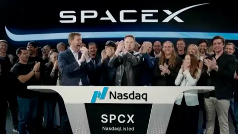 Elon Musk wearing a black leather jacket, stands in the center of crowds and in front of a SpaceX sign.