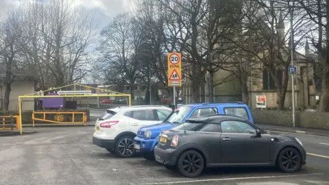 Three cars are parked in a car park next to a road with a school on it.