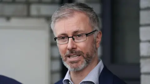 Roderic O'Gorman - a man with grey hair wearing a white and blue collared shirt, navy jacket and black rectangle framed glasses.