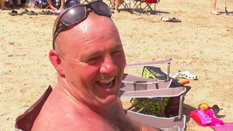 A sunburnt man sits in a chair on the beach. He is bald, topless and has sunglasses on his head. 