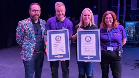 BBC A row of four people on a theatre stage. A man with a beard and a multi-coloured paint-splash suit jacket stands next to a man in a blue t-shirt holding a framed certificate. Next to him is a blonde haired woman in a blue t-shirt. She is holding a framed certificate and next to her is a red haired woman in a blue t-shirt.