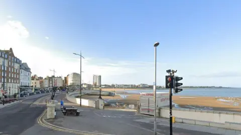 Google The sea and beach in Margate seen from the road. The steps leading down to the beach are seen in the distance. The sky is blue and there are buildings lining the road next to the beach.