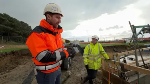 BBC Tom Le Gallais at the site wearing a orange hi-vis jacket and a cream hard hit. Another man is also at the site wearing a yellow hi-vis jacket and safety helmet. The site is muddy.