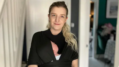 A woman with blonde hair is wearing a black baby sling. You can just see the side of the baby’s head inside the sling asleep.