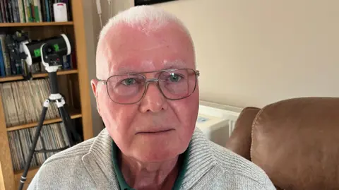 John Devine/BBC Alan Florence has short white hair. He is sitting on a tan coloured leather armchair. He is wearing silver framed glasses, a green shirt and a beige jumper. Behind him is a bookshelf with records on it.