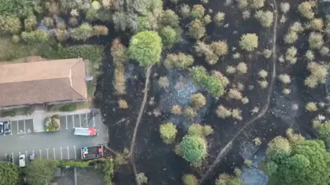 An aerial image of an area of land blackened after a fire, with some trees remaining green and having seemingly survived the blaze. There is a fire engine in a car park to the left hand side of the image, with a building and some other parked vehicles alongside.