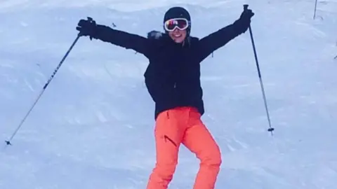 Becky Williams Becky Williams on snow in ski clothes and helmet, holding ski poles aloft