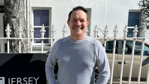BBC Dr Christopher Pich. He has short brown hair and is wearing a grey jumper. He is stood in front of some white railings. 