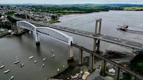 An aerial view of the River Tamar showing both the railway bridge and the road crossing with boats in the water and views of field in the distance.
