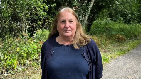 Elaine Young has straight blonde hair and wears navy blue top and cardigan. The path which is what is left of the former Penshaw to Sunderland railway is behind her.