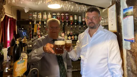 Two men are standing at a bar inside a pub. They are both holding up glass cups with beer in them. The man on the left has grey hair and glasses, and the man on the right has dark hair with grey facial hair. There are bottles in the background.