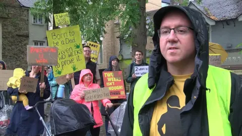 A man wearing a waterproof jacket gives an interview. Behind him, women and men are striking for better paternity leave. They hold placards with slogans such as "2 weeks? What a dad joke" and "Parenting is work". It is a rainy day.