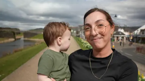 A woman with tied-back brown hair and glasses, wearing a black t-shirt and gold necklace, holding a toddler in a green t-shirt who is looking away from the camera. They are stood on a path with a town centre and a canal behind them