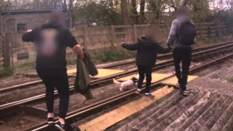 Two adults walking along tracks with a child and a small white dog. The adult at the back is carrying their jacket, while the adult at the front is holding the child's hand. The child is holding the dog's lead.