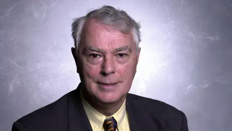 BBC A man with short grey hair, looking at the camera. He is pictured from the chest up. He is wearing a yellow shirt, a brown tie and a dark grey suit. 