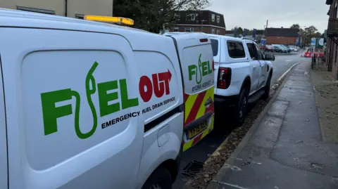 A white van that says "Fuel Out" on its side. It has a back door open next to a white truck parked on a residential street.