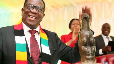 Emmerson Mnangagwa wears a scarf bearing Zimbabwe's national colours. He smiles next to one of the stone bird sculptures.
