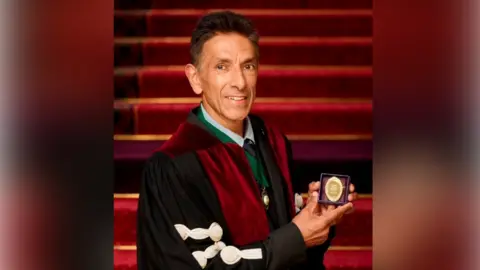 A head and shoulders shot of Professor Sunil Bhandari, wearing a black and red robe, holding a small gold medal which sits in a box. He has brown hair and is smiling at the camera. He is stood in front of a set of red-carpeted stairs.