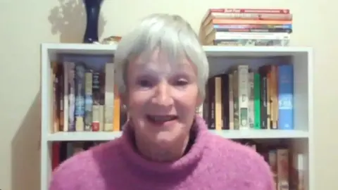 Leanne Seignior photographed in a room with a bookshelf behind her. She has grey short hair and is wearing a thick pink turtle neck jumper.