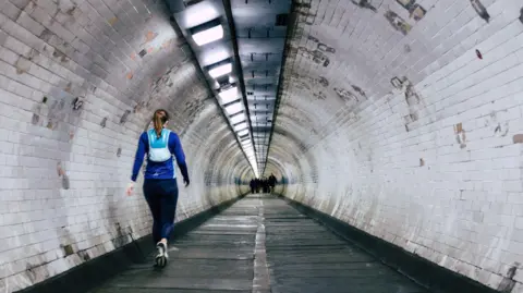 A woman in running gear walks through a long, tiled pedestrian tunnel, with bright overhead lights and a small group of people visible in the distance.
