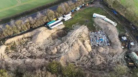 Shaun Whitmore/BBC Aerial image shows one uncovered waste pit, and a large area of mounds and flattened earth, suggesting others have been dug and covered over. There is a grassy area, solar panels and several freight containers. A road runs across the top of the picture, and another track can also be seen on the right of the frame.