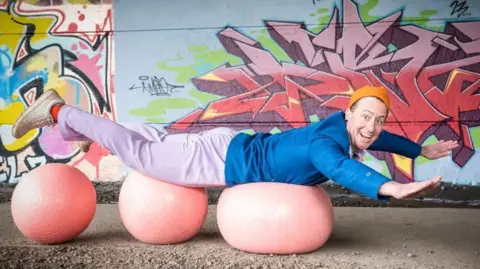 A man in a blue jacket and lilac trousers and party hat smiles at the camera as he lays across and balances on three medicine balls.