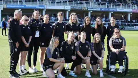Katherine Selby Sharland and her teammates on the Hereford FC pitch. The players are lined up for a team photo in match black and grey kits. Sharland is on the end and sat in her wheelchair.