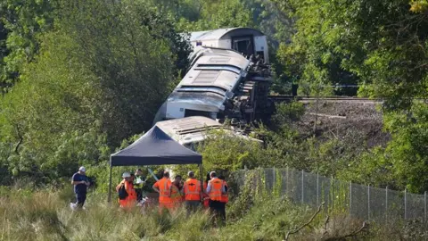 PA Media Three train carriages are derailed on a railway embankment near Lewes. Several people in orange high-visibility clothing stand beneath a temporary canopy in the foreground while the damaged carriages remain among trees and vegetation beside the track.
Show more lines