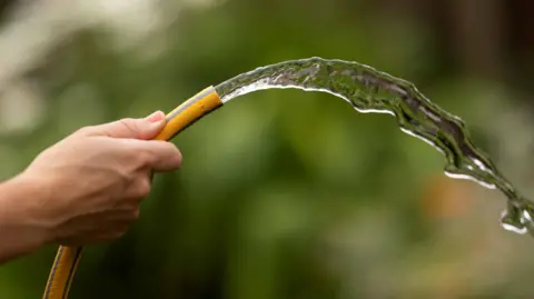 A man's hand holding a yellow hose pipe with water flowing out of it.