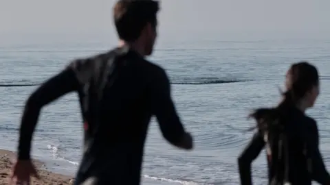 Two swimmers about to enter into the sea in wetsuits. 