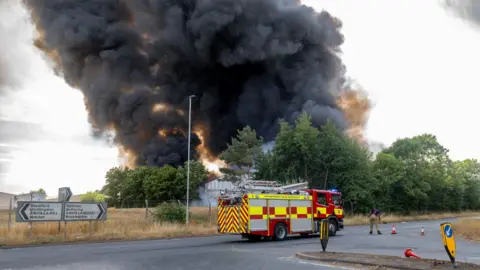 A fire engine parked on the road and there is a large plume or grey/black smoke behind it 