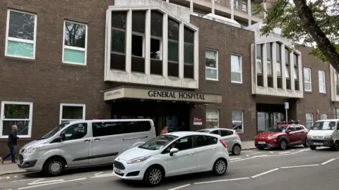 Cars drive past a brick building with the words General Hospital above an entrance. Passers by are walking in the street.