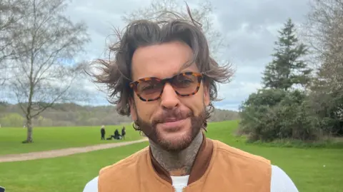 Alicia Owen/BBC Pete Wicks has a big floppy brown fringe which is windswept, thick framed glasses and is wearing a brown vest jacket. Behind him is the grass lawns of the country park and trees.
