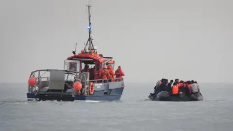 A French rescue boat with crew members in orange uniforms and life jackets escorts a small, overcrowded inflatable boat. The inflatable boat is filled with people wearing life jackets, believed to be migrants, as it moves away from the French coast.