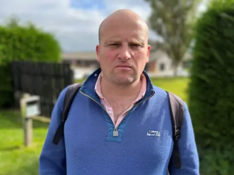 Peter Todd wearing a blue jumper standing outside New Craigs Hospital in Inverness, which is in the background between a fence and a hedge