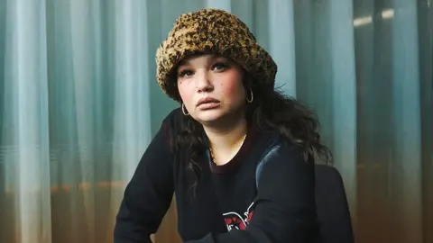 BBC / Jonathan Birch Lola Young wearing a black long sleeved top, looking into the camera, wearing a printed winter hat, with light blue coloured curtains behind her.