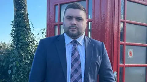 Alan Lewis in front of a red phone box. He has dark hair and beard, wearing a dark blue blazer, light shirt and check tie. A telephone pole covered in ivy can be seen to his right.