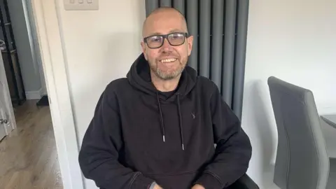 Family Handout Nick Ball wearing a black hoodie and black framed glasses. He has a bald head and a closely shaven beard. He is sitting down in a white living room beside a grey dining chair. He is smiling at the camera, but looks weak from cancer treatment.