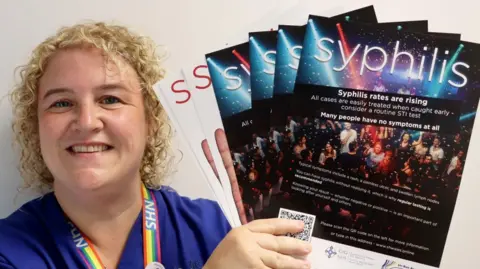 Swansea Bay University Health Board Carly is holding up posters advertising testing for syphilis. She is wearing dark purple medical scrubs and a rainbow NHS lanyard around her neck. She has short curly blonde hair and is smiling at the camera.