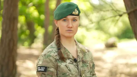A woman in a military uniform and wearing a beret is standing outdoors under trees. She is looking in the direction of the camera. The background behind her is blurred. 