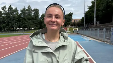 Head and shoulders image of wheelchair racer Melanie Woods. She is on running track and is facing into the camera. She is white and has brown hair tied back and black sunglasses on the top of her head. She's wearing a light khaki green rain jacket and a grey t-shirt and has a gold necklace and gold earrings