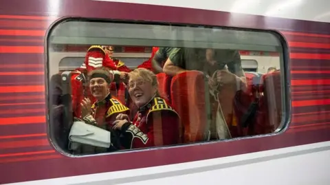 PA Media Some guards through the train window. Two of them are smiling into the camera while sat on train seats. They are not wearing their hats and people around them are stood up.